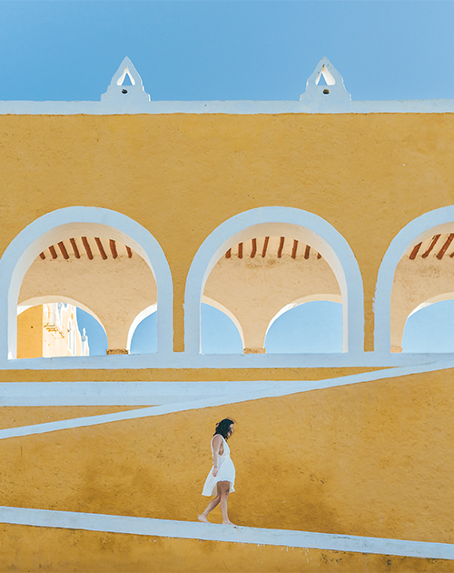 A woman in white dress walking through the vibrant yellow and white arches of the colonial architecture in Yucatan, Mexico